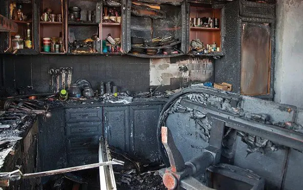 A severely fire-damaged kitchen with cabinets, worktops and appliances burned through and coated in soot.